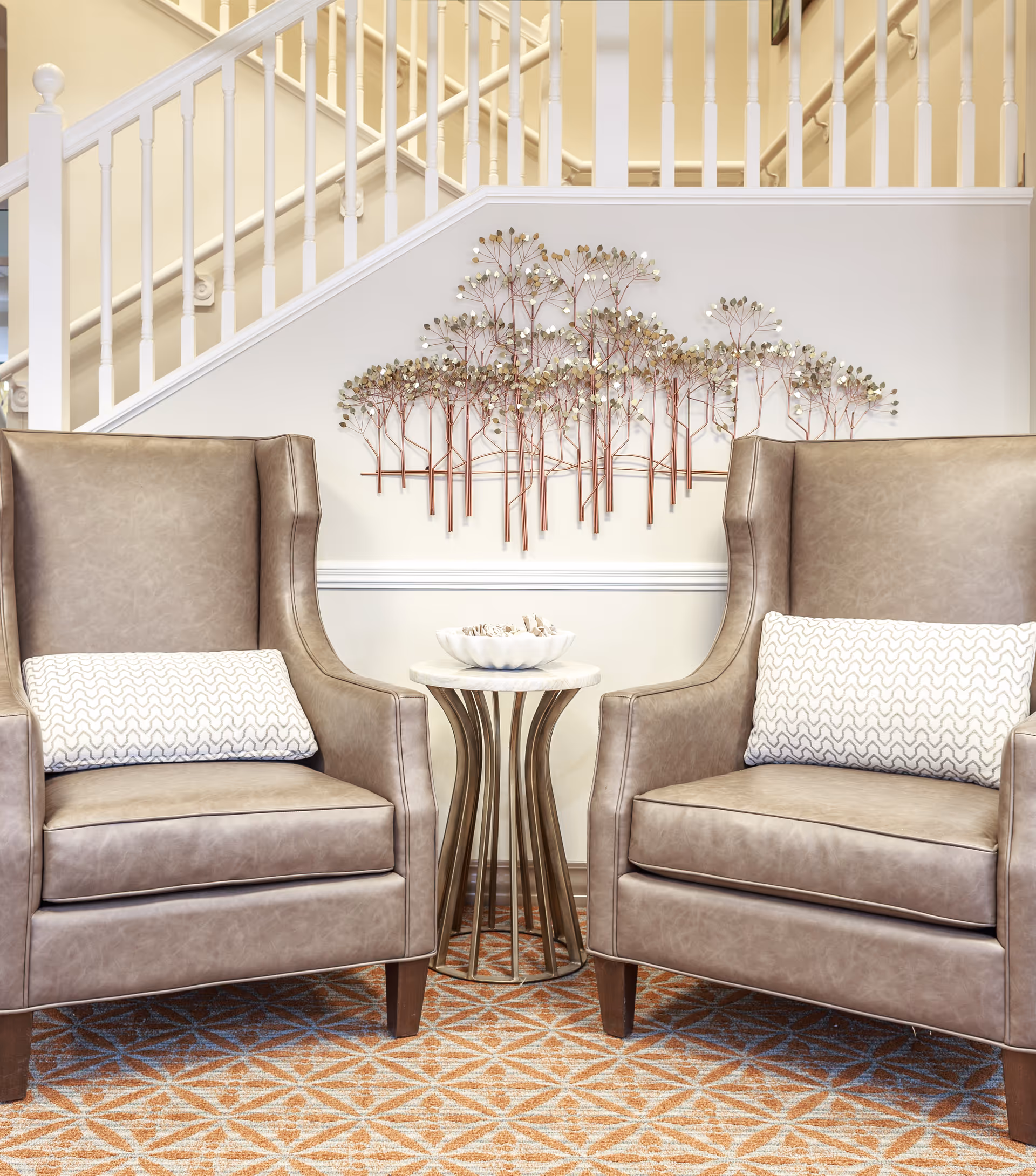 Two beige leather armchairs with patterned cushions are positioned on a geometric patterned carpet in front of a white staircase railing. Between the chairs is a small round side table with a decorative bowl on top. A metal wall art piece depicting trees is mounted on the wall behind the table.