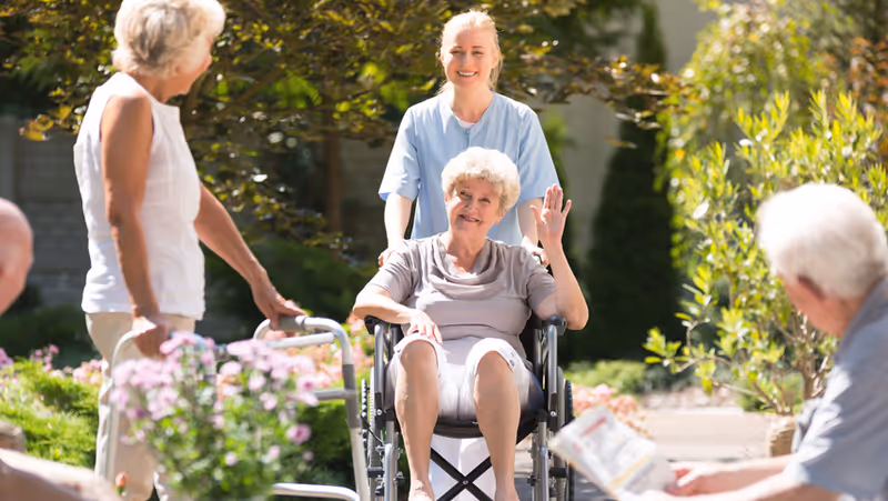 An elderly woman in a wheelchair waving and smiling outdoors, accompanied by a caregiver standing behind her. Two other elderly individuals are nearby, one with a walker and the other reading a newspaper, surrounded by greenery and flowers in a garden setting.