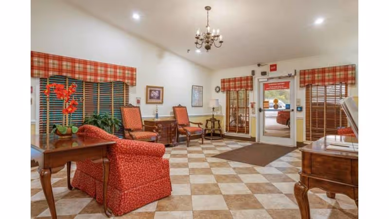 A cozy waiting area with red patterned armchairs and wooden furniture. The floor has a checkered tile pattern in beige and brown. Windows and the entrance door have wooden blinds and red plaid valances. A chandelier hangs from the ceiling, and there are framed pictures on the walls.