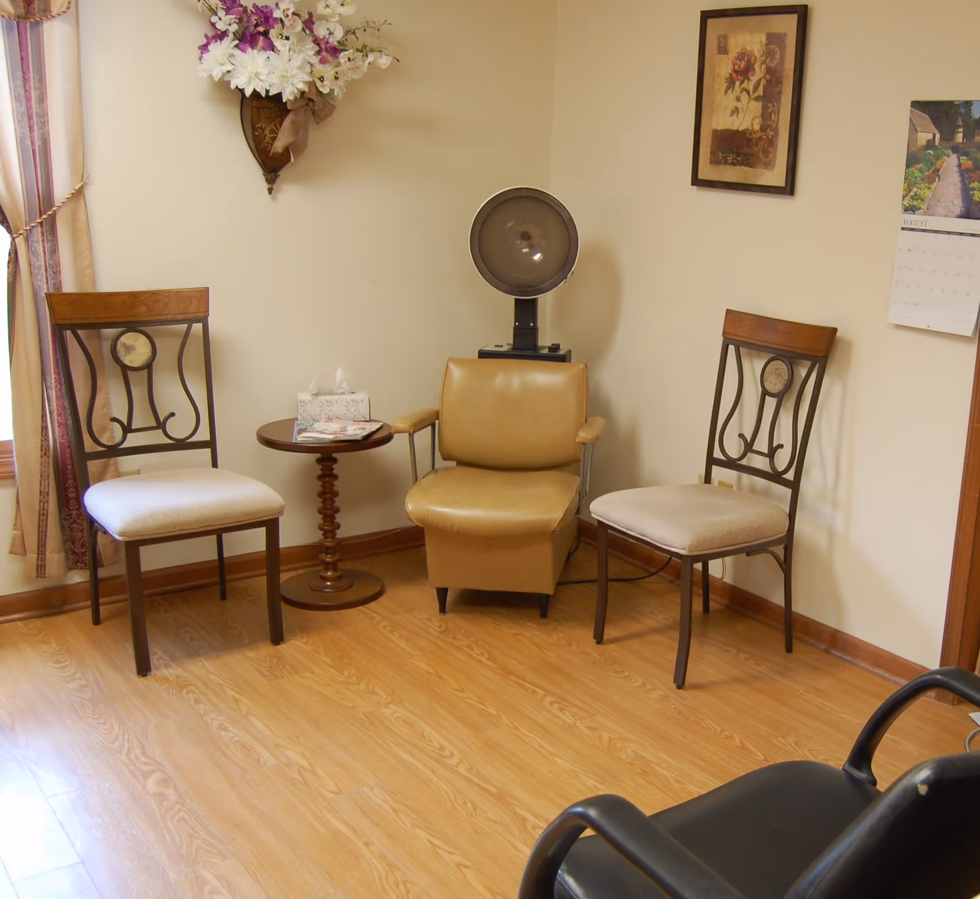 A small seating area with two wooden chairs with cushioned seats, a tan leather chair with armrests, and a round wooden side table holding a tissue box and some magazines. A vintage hair dryer is positioned behind the leather chair. The room has light-colored walls, a wooden floor, a floral wall decoration, a framed picture, and a calendar on the wall.