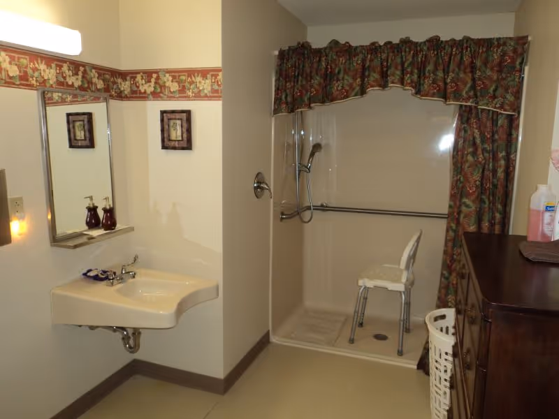 A bathroom with a wall-mounted sink and mirror on the left side. There is a shower area with a handheld showerhead, grab bars, and a white shower chair. The shower has a floral curtain valance. On the right side, there is a dark wooden dresser with a bottle of hand soap and a laundry basket next to it. The walls have a floral wallpaper border near the ceiling.