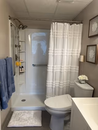 A clean and well-lit bathroom featuring a white shower with a curtain, a toilet, a white bath mat on the floor, a towel rack with blue towels, and a small vase with white flowers on the toilet tank. Two framed floral artworks hang on the wall above the toilet.