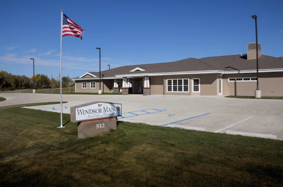 One-story assisted living building with a paved front parking area, an American flag, and a lawn sign reading "Windsor Manor".