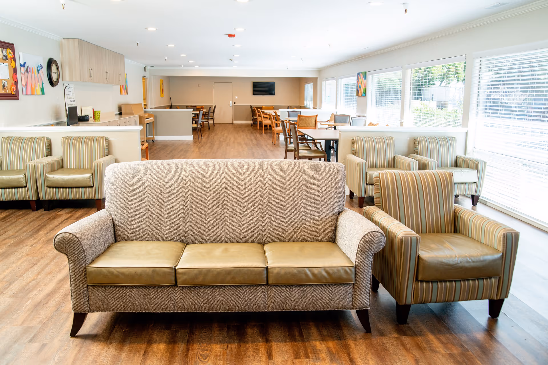 A bright and spacious common area in an assisted living facility featuring a beige sofa with leather cushions and a matching striped armchair in the foreground. The room has large windows with blinds on the right side, letting in natural light. In the background, there are multiple tables and chairs arranged for dining or activities, with colorful artwork on the walls and a television mounted on the far wall.