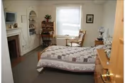 Furnished bedroom with a bed covered by a patchwork quilt, dresser, fireplace with built-in shelves, and a desk and chair by a window.