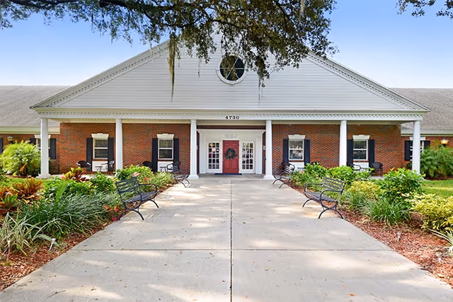 Front exterior view of a single-story brick building with white columns and a triangular pediment. The entrance has double doors with a wreath, and there are benches and landscaped greenery along the concrete walkway leading to the entrance. The building number 4730 is displayed above the entrance.