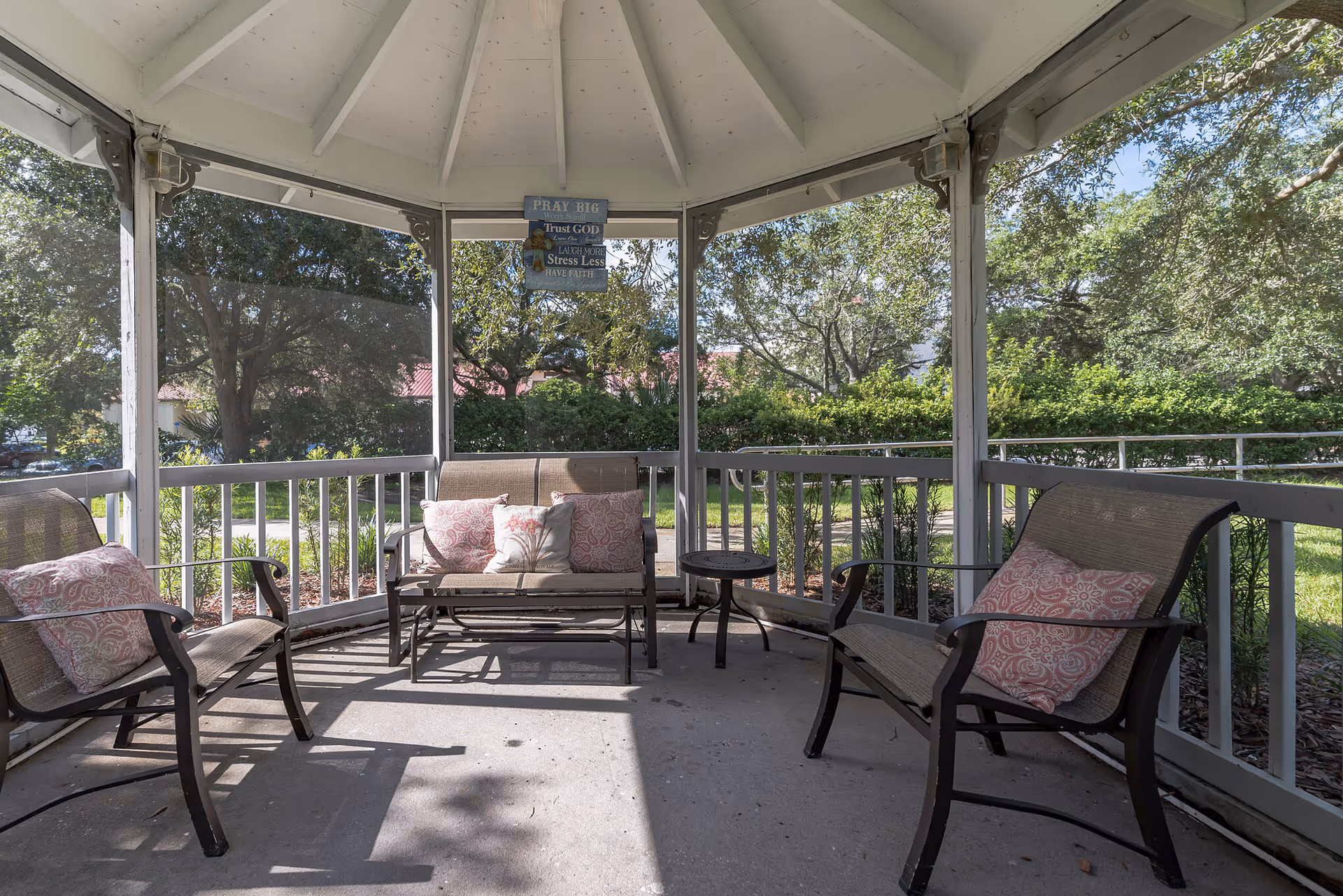 A screened-in outdoor gazebo with three metal-framed benches with mesh seats and backs, each with patterned cushions. There is a small round table in the center. The gazebo is surrounded by greenery and trees, with sunlight casting shadows on the concrete floor. A decorative sign hangs from the ceiling with the words 'PRAY BIG, Trust GOD, Stress Less, HAVE FAITH.'