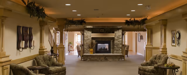 Warmly lit communal lounge with a central stone fireplace, upholstered chairs, columns, and decorative plants.