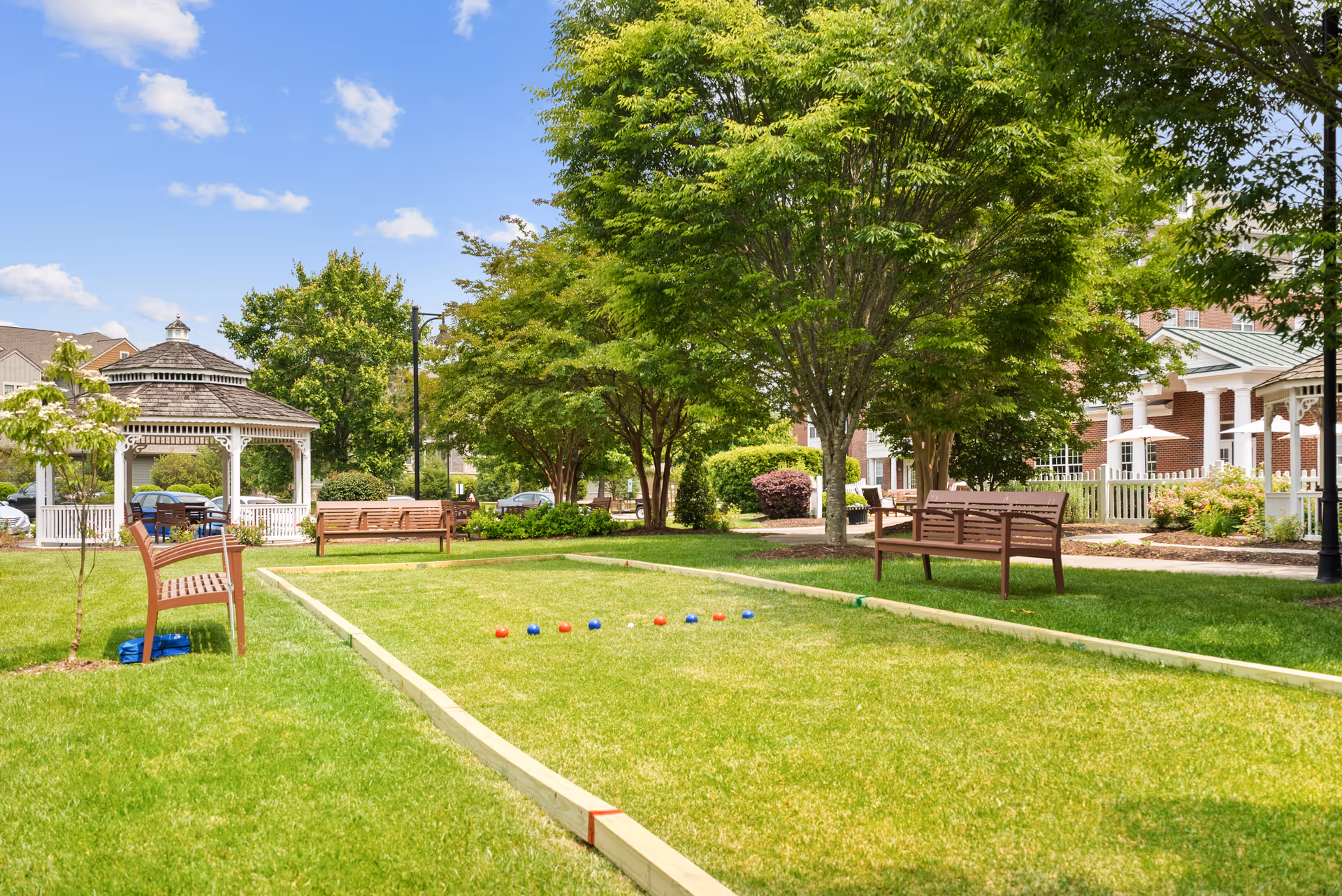 Outdoor bocce ball court with wooden benches on a grassy area surrounded by trees and shrubs. A white gazebo is visible in the background along with a brick building and a clear blue sky with some clouds.