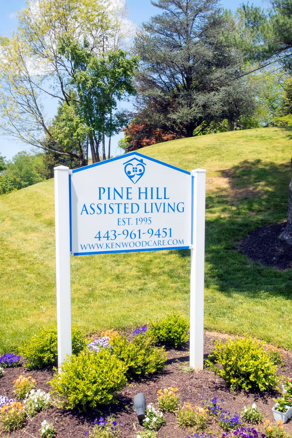 Outdoor image of a white and blue sign for Pine Hill Assisted Living, established in 1995, with contact number 443-961-9451 and website www.kenwoodcare.com, surrounded by green grass, bushes, flowers, and trees in the background.