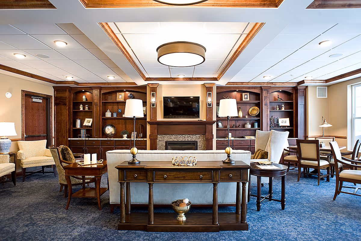 A spacious and well-lit living room area with a blue carpet and wooden furniture. The room features a central fireplace with a mounted flat-screen TV above it, flanked by built-in wooden shelves filled with decorative items. There are multiple seating options including armchairs, a sofa, and dining chairs around tables. Two table lamps are placed on a wooden console table behind the sofa. Large windows on the right side allow natural light to brighten the room.