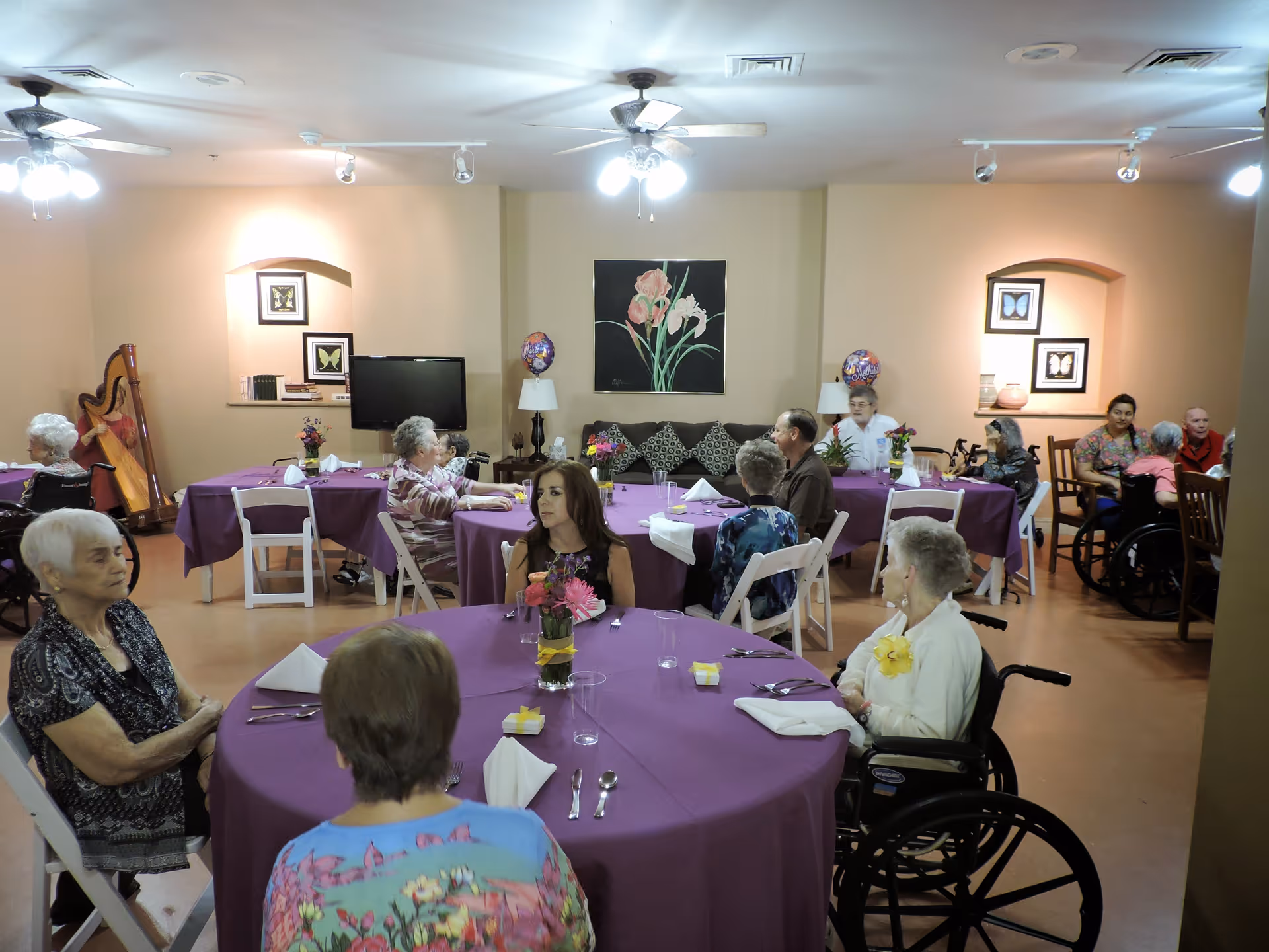 Seniors seated around round tables with purple tablecloths in a dining/activity room at an assisted living facility.