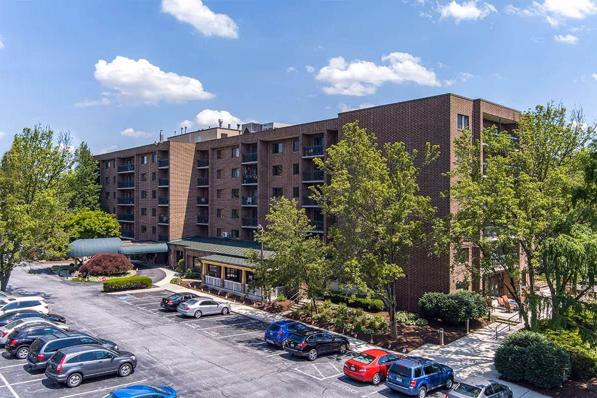 Exterior view of a multi-story brick senior living building with a parking lot, trees, and an entrance canopy.