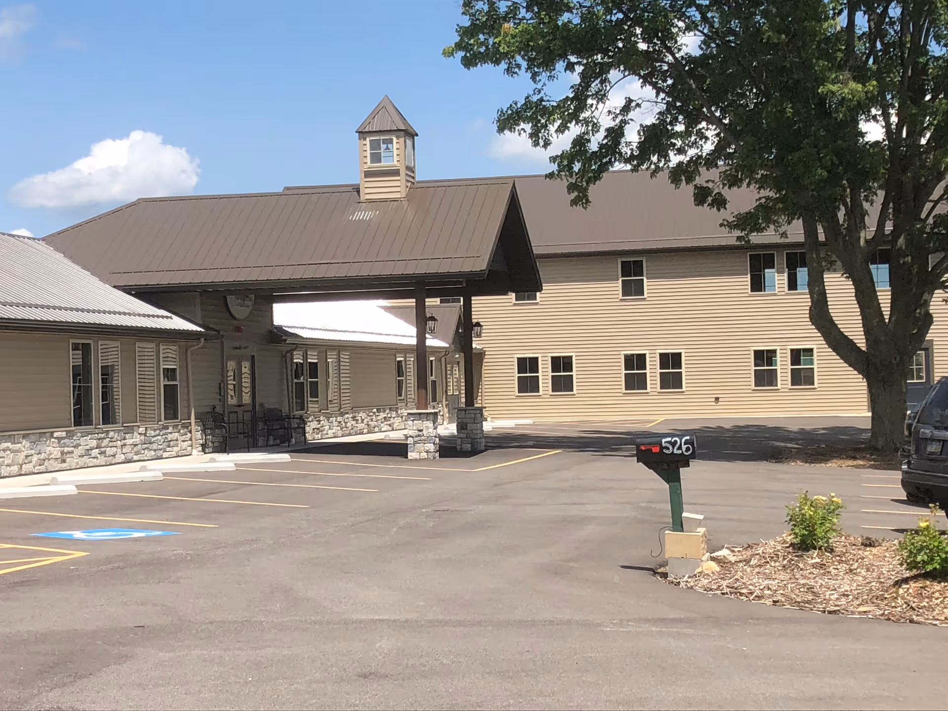 Parking lot and entrance canopy of a beige senior living building with a mailbox labeled 526 and a large tree.