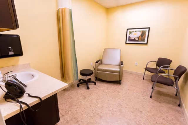 A small room with beige walls and flooring, featuring a sink with a countertop and a telephone on the left side. There is a beige recliner chair, a small black stool, and two brown chairs with armrests. A framed picture of a white flower hangs on the wall.