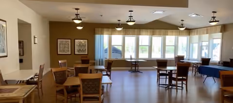 A spacious dining room with multiple wooden tables and chairs arranged neatly. Large windows along one wall allow natural light to fill the room. Ceiling lights hang evenly spaced across the ceiling. The room has light-colored walls and a wooden floor.