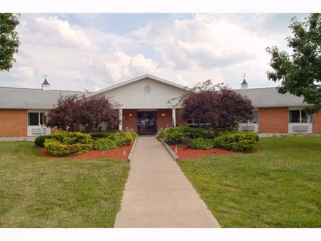 Front entrance of a single-story brick senior living building with a central walkway, shrubs, and trees.