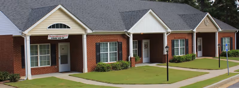 Exterior view of a single-story brick building with white trim and multiple entrances, each with a small porch and windows with black shutters. There is a sign above one entrance that reads 'This Unit OPEN FOR INSPECTION COME ON IN.' The building is surrounded by neatly trimmed grass and paved walkways.