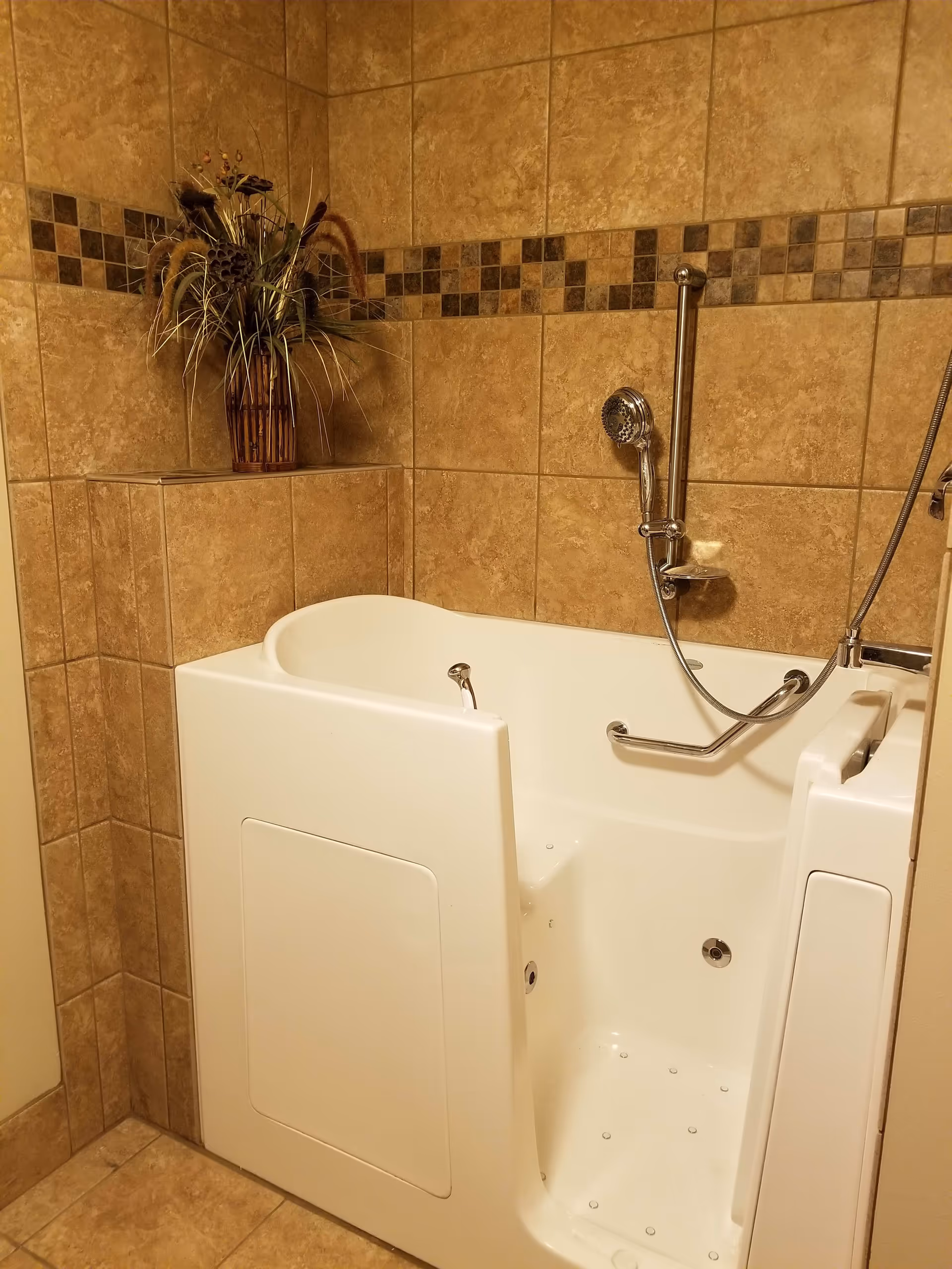 A walk-in bathtub with a door for easy access, surrounded by beige tiled walls with a decorative tile border. There is a handheld showerhead and a grab bar inside the tub. A vase with dried plants is placed on a tiled ledge in the corner.