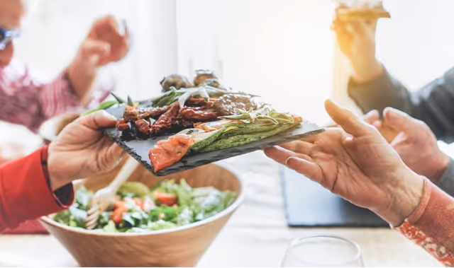 Several people pass a slate platter of appetizers over a salad bowl at a dining table.