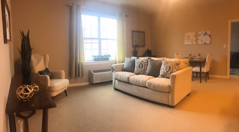 A cozy living room with a beige sofa adorned with patterned and solid cushions, a beige armchair with a throw blanket and pillow, a wooden console table with decorative items, a window with curtains letting in natural light, and a small dining area with a table set for two in the background.