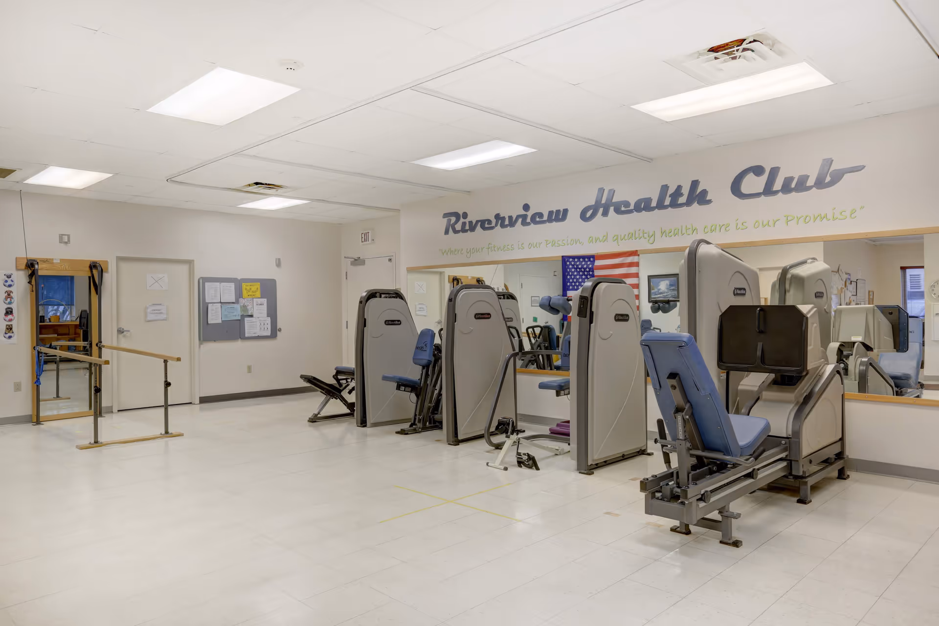 Interior view of Riverview Health Club fitness room with exercise machines, a ballet barre, mirrors on the wall, and an American flag. The room has white walls, a tiled floor, and fluorescent ceiling lights.