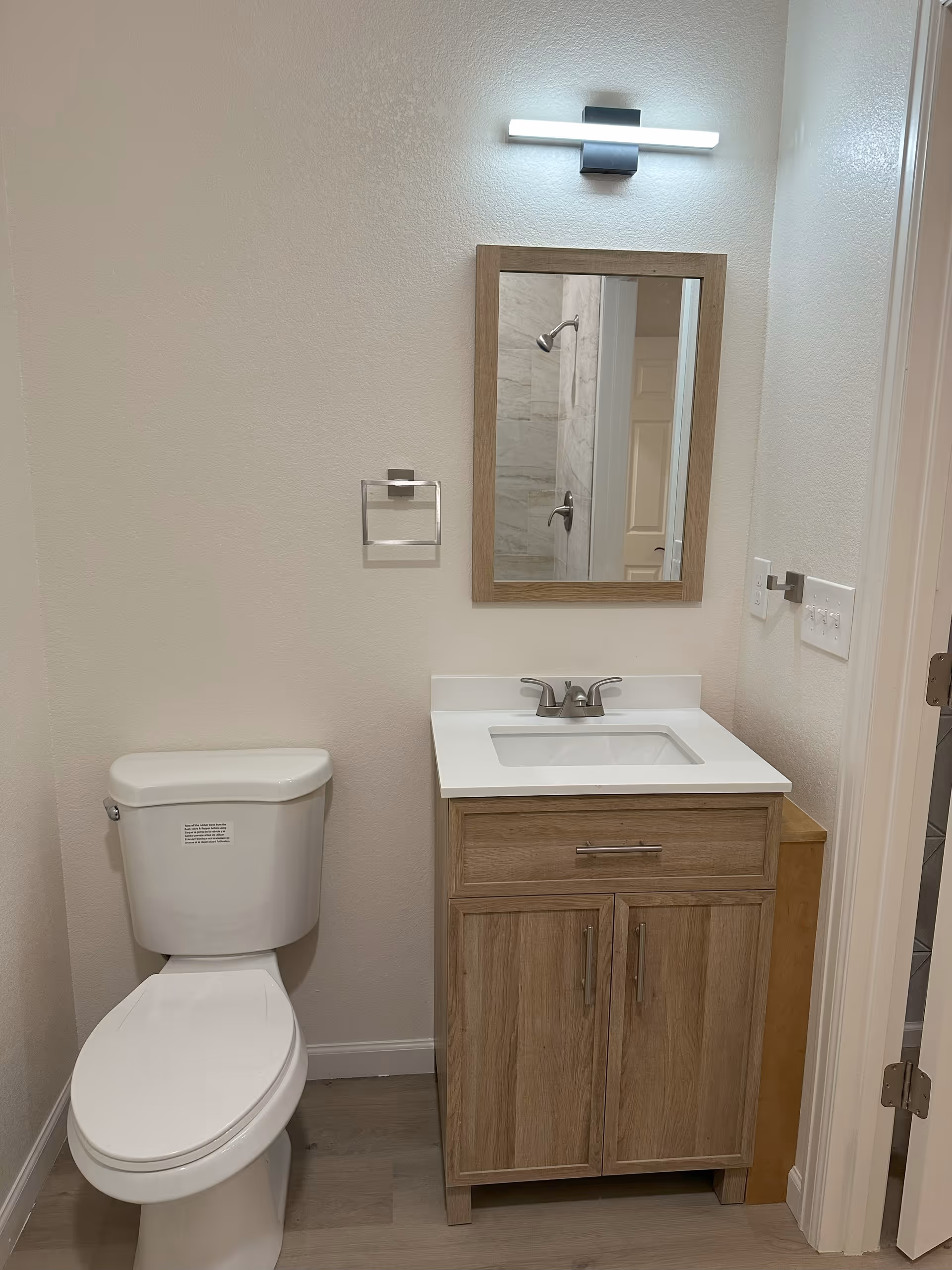 Compact bathroom with a toilet, wooden vanity with sink and mirror, and a wall-mounted light above.