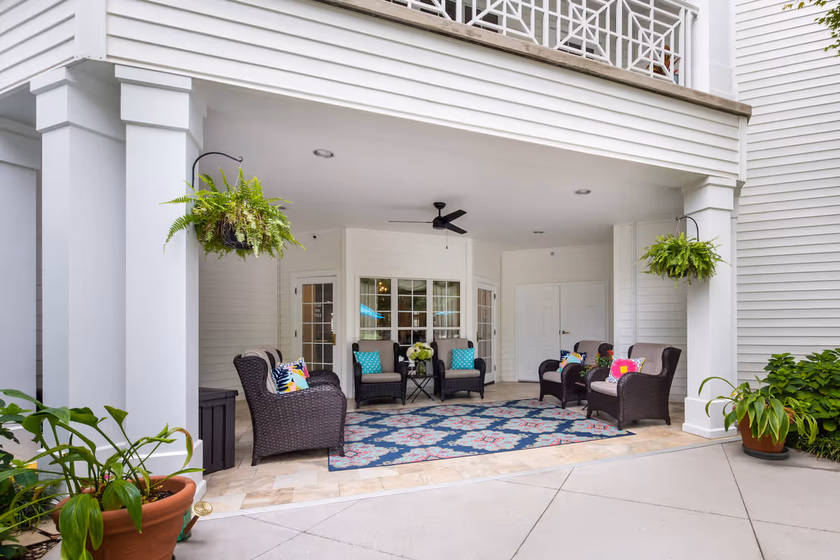 Covered outdoor seating area with wicker armchairs featuring colorful cushions arranged around a patterned blue and pink rug. Two hanging green plants are suspended from the ceiling, and potted plants are placed on the ground. The area is part of a white building with large windows and double doors, and a ceiling fan is mounted overhead.