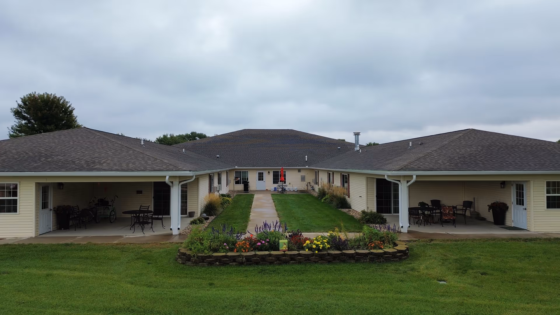 View of a single-story senior living facility forming a U-shaped exterior courtyard with a central walkway, lawn, and flowerbed.
