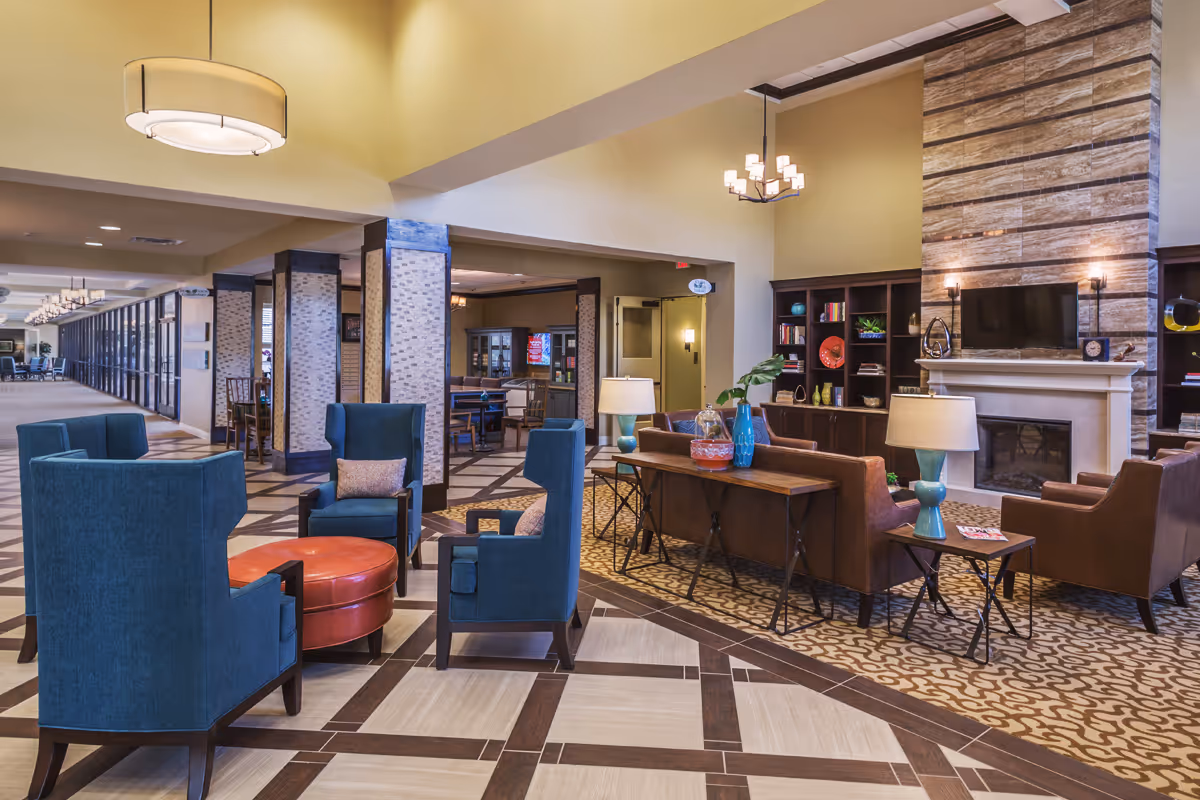 A spacious and well-lit common area in a senior living facility featuring a seating arrangement with blue armchairs around a round leather ottoman, a brown leather sofa set near a fireplace with a mounted TV above it, and built-in bookshelves with decorative items. The area has patterned carpet and tile flooring, modern hanging light fixtures, and a long hallway with large windows on one side.