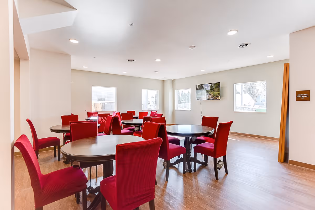 A bright room with multiple round tables surrounded by red upholstered chairs. The room has light-colored walls, wooden flooring, several windows letting in natural light, and a wall-mounted television. A sign on the wall indicates a maximum occupancy of 35.