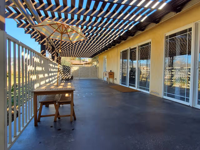 Covered outdoor patio with a wooden pergola, umbrella-shaded table and benches, and sliding glass doors along the building.