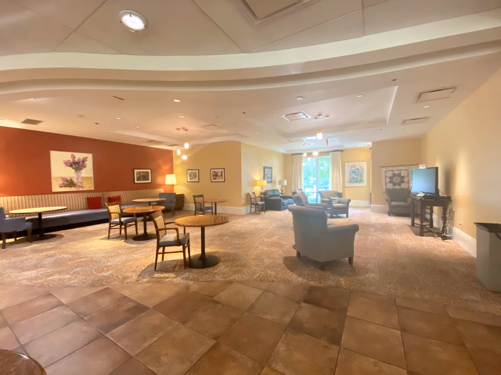 A spacious common area with a mix of seating including armchairs, small round tables with chairs, and a long bench against a red accent wall. The room has warm lighting, carpeted flooring with a patterned design, and a tiled area in the foreground. There are framed pictures on the walls, a TV on a stand, and large windows with curtains letting in natural light.