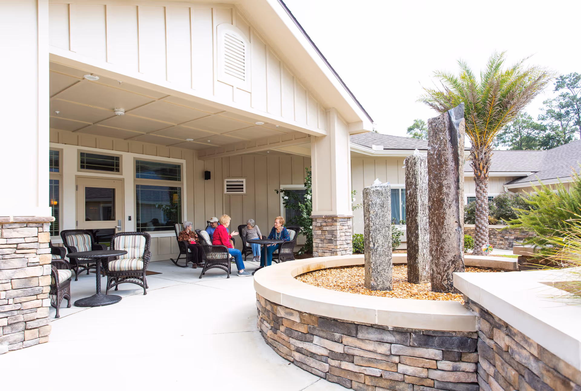 Outdoor covered patio area at Azalea Gardens Assisted Living & Memory Care with several elderly people sitting and chatting around a table. The patio features stone pillars and comfortable wicker chairs with cushions. In the foreground, there is a circular stone planter with tall stone sculptures and a palm tree nearby.