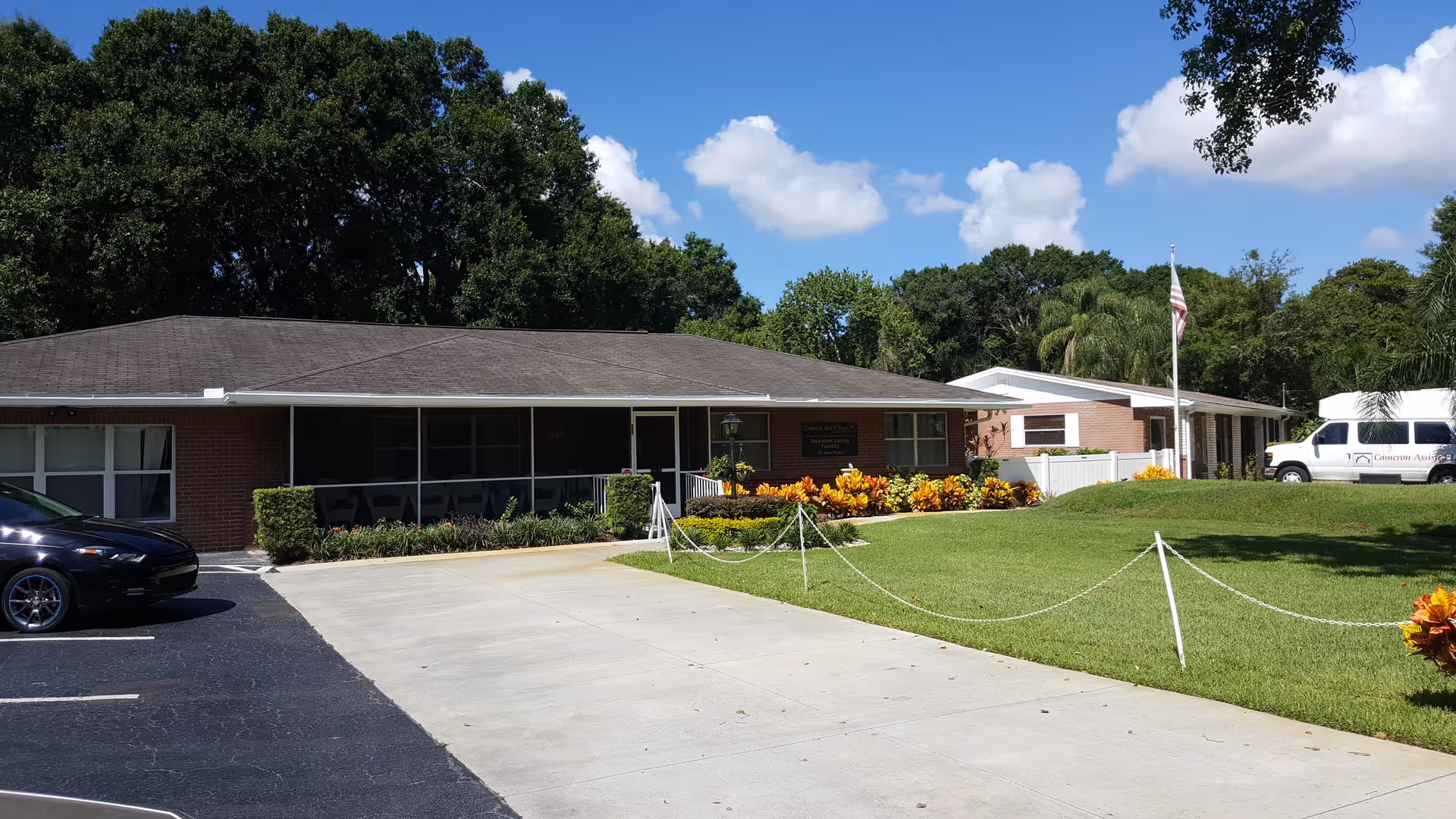 Front exterior of a single-story assisted living building with a driveway, lawn, American flag, and parked vehicles.