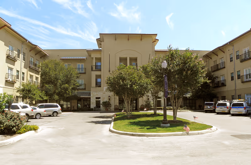 Front exterior view of a three-story assisted living facility building with a driveway, parked cars, trees, and a lamppost in the center of a circular landscaped area under a clear blue sky.