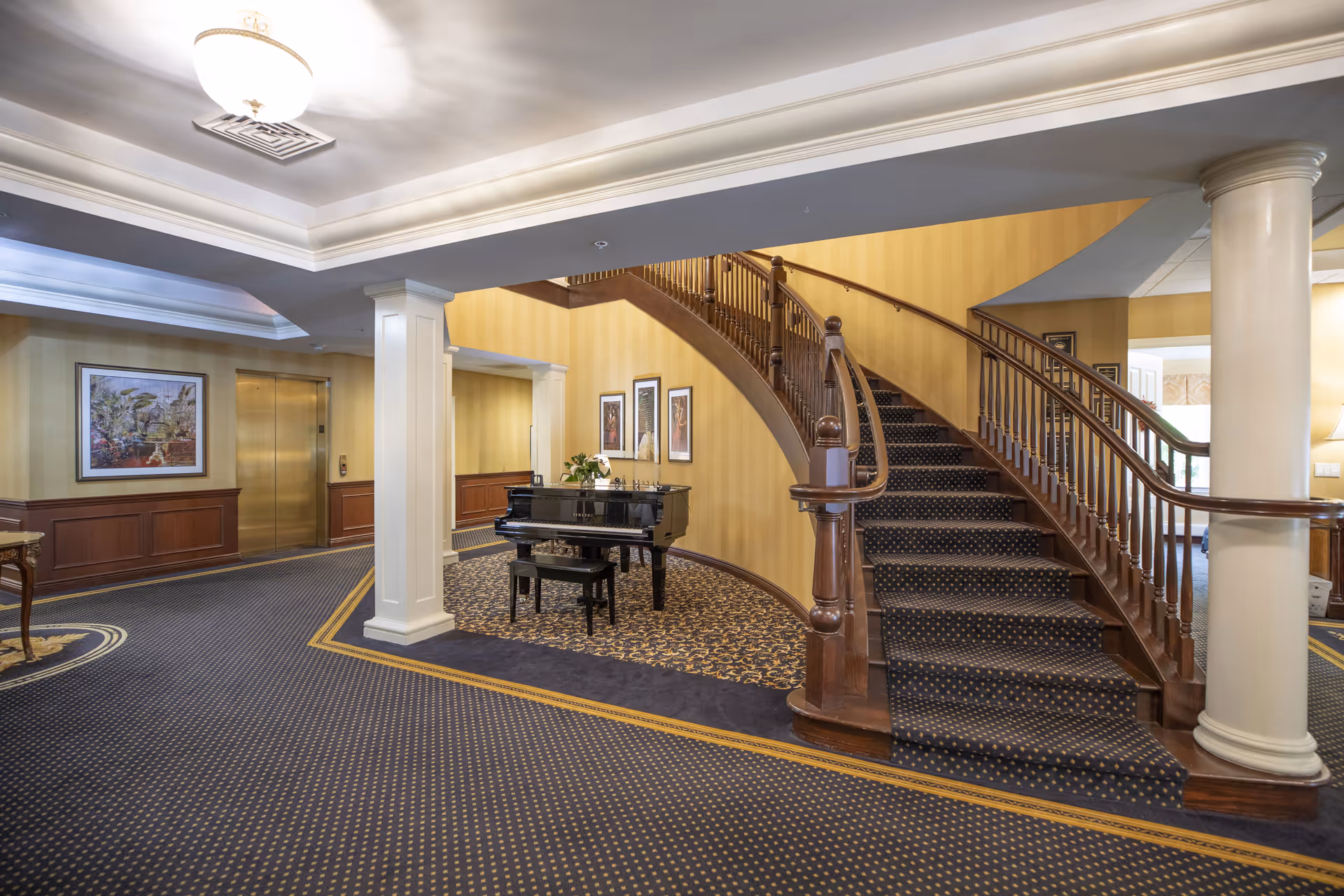 Lobby interior with a curved wooden staircase, a black grand piano, columns, and patterned carpeting.