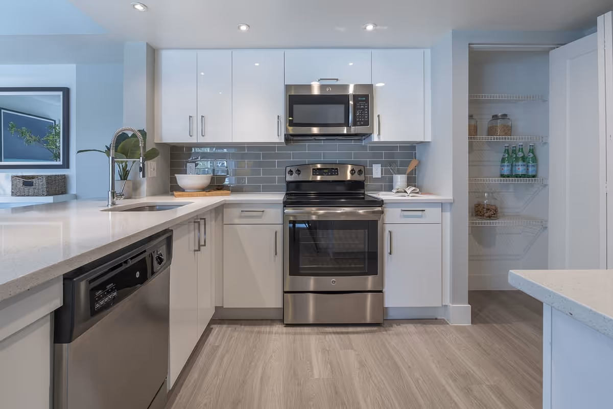 Modern kitchen with white cabinets, stainless steel appliances including a stove, microwave, and dishwasher. There is a sink with a tall faucet on the left side and an open pantry with shelves holding bottles and jars on the right. The backsplash is made of gray subway tiles and the floor is light wood.