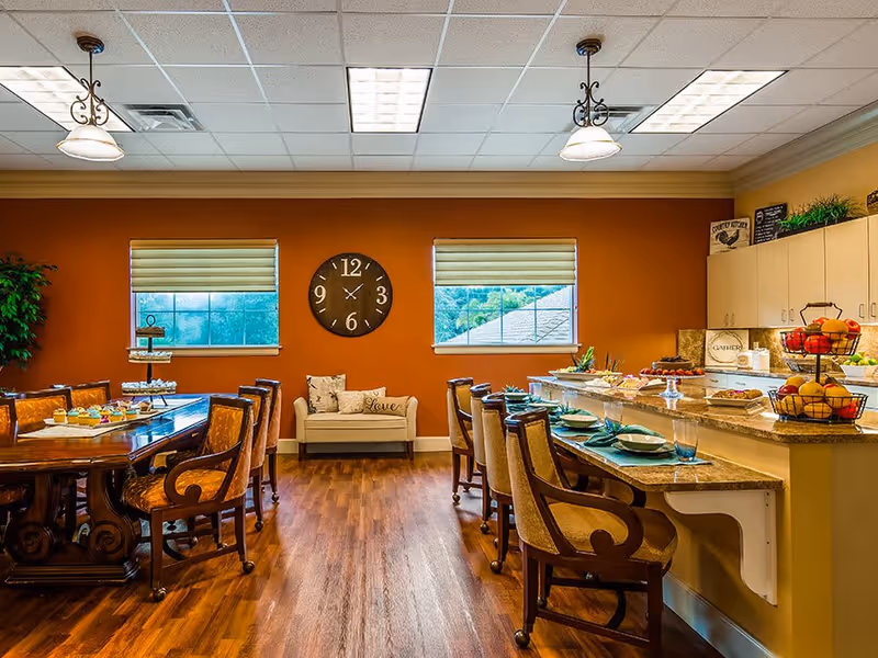 A warm dining area with wooden floors and orange walls featuring two windows with blinds and a large round clock between them. There is a wooden dining table with cushioned chairs on the left side and a counter with bar stools on the right side. The counter is set with plates, glasses, and various food items including fruit baskets and desserts. A small cushioned bench with pillows is placed against the wall under the clock. Ceiling lights illuminate the room.