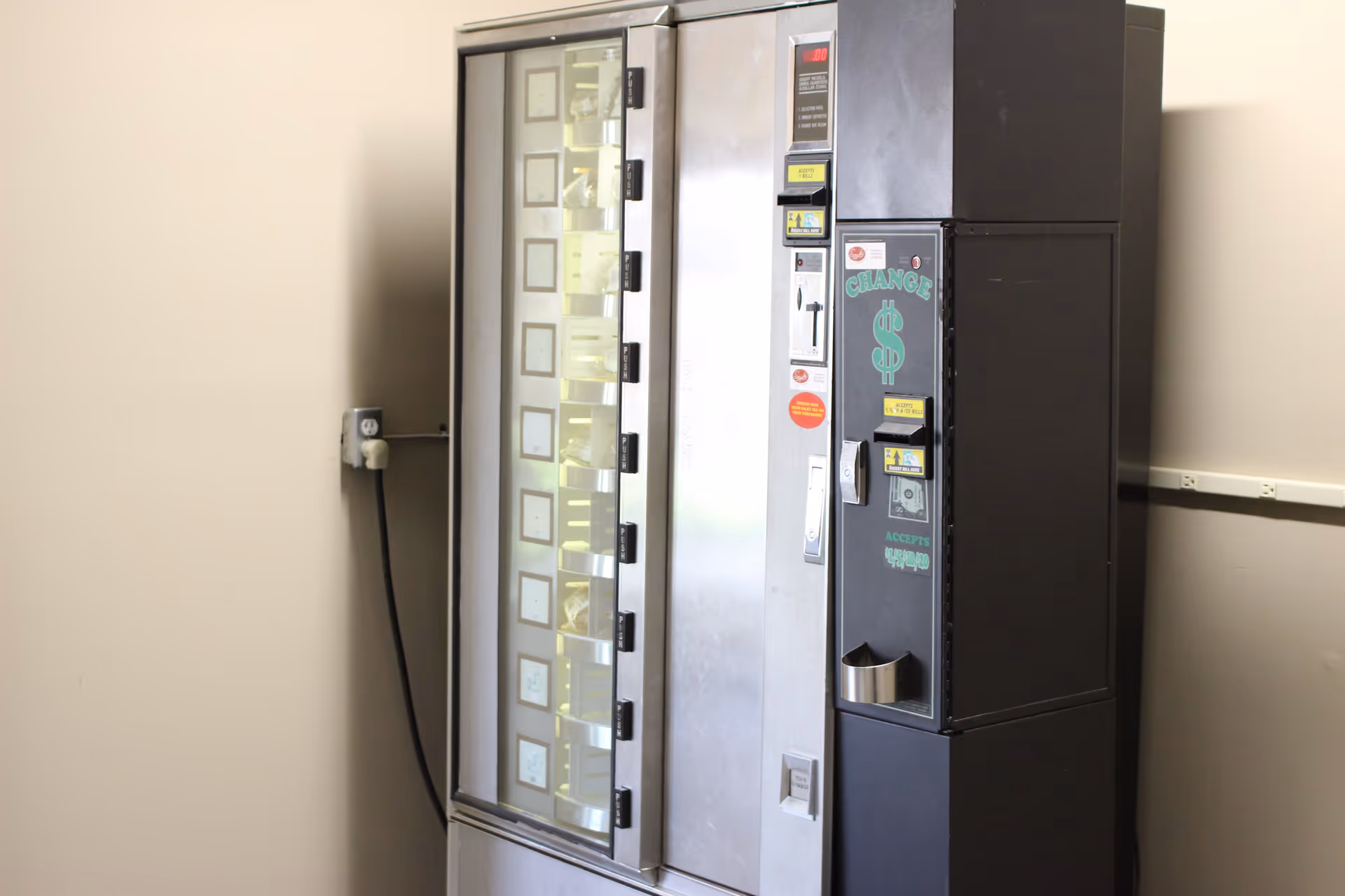 A silver vending machine with an attached change machine standing against a beige wall.