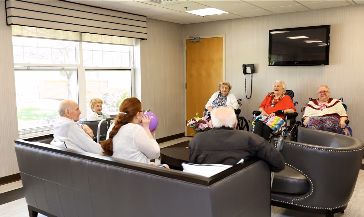 A group of elderly people sitting and socializing in a well-lit common area with large windows. Some are seated on a black leather couch, while others are in wheelchairs. A flat-screen TV is mounted on the wall above a phone, and a wooden door is visible in the background.