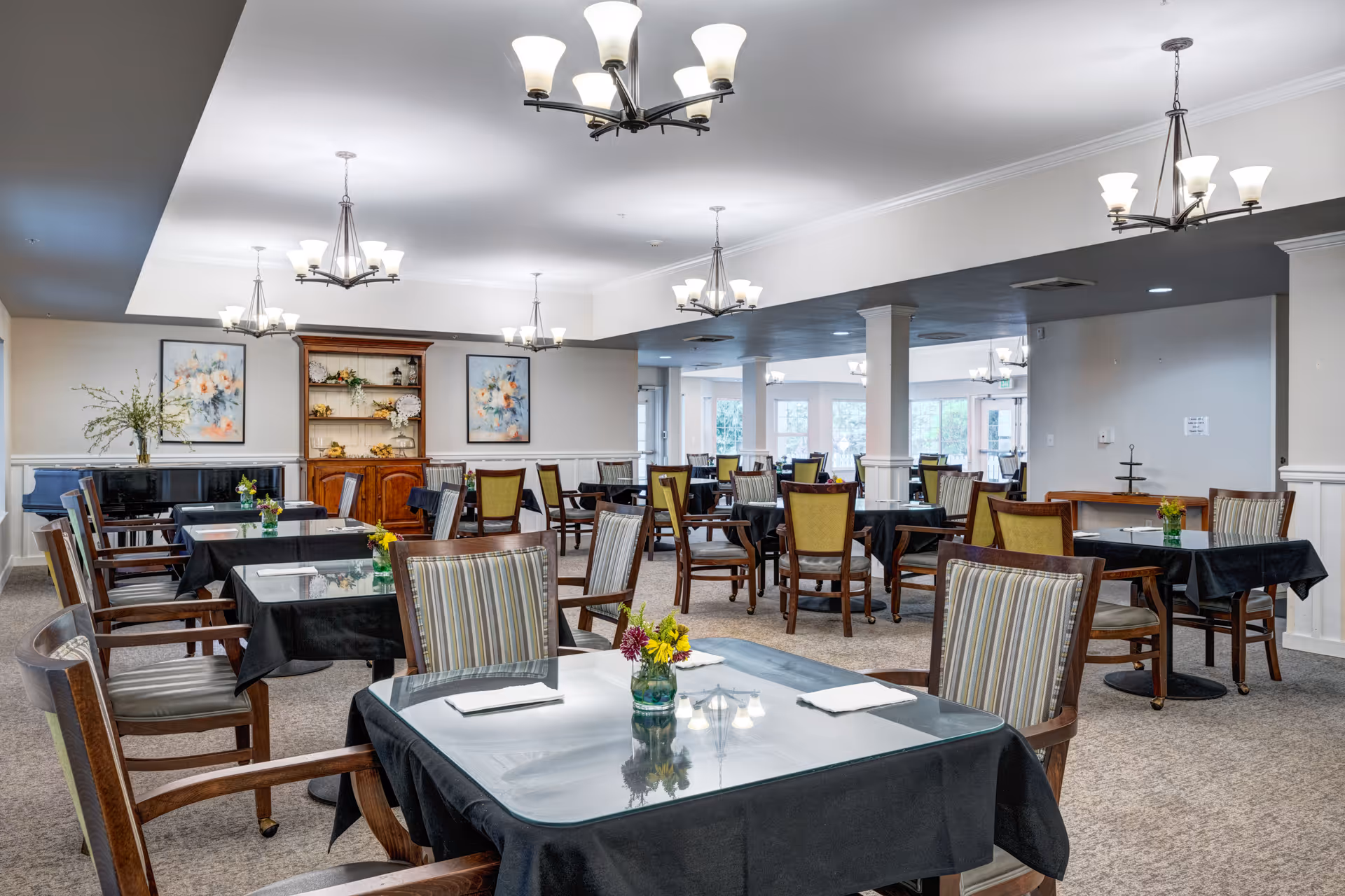 A spacious dining room with multiple tables covered in black tablecloths, each set with napkins and small flower arrangements. The room features wooden chairs with striped and solid upholstery, chandeliers hanging from the ceiling, framed floral artwork on the walls, and a wooden cabinet with decorative items. Large windows in the background allow natural light to fill the space.
