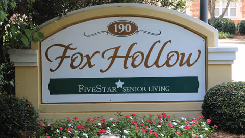Outdoor sign for Fox Hollow Senior Living Community with the address number 190, surrounded by greenery and flowers.