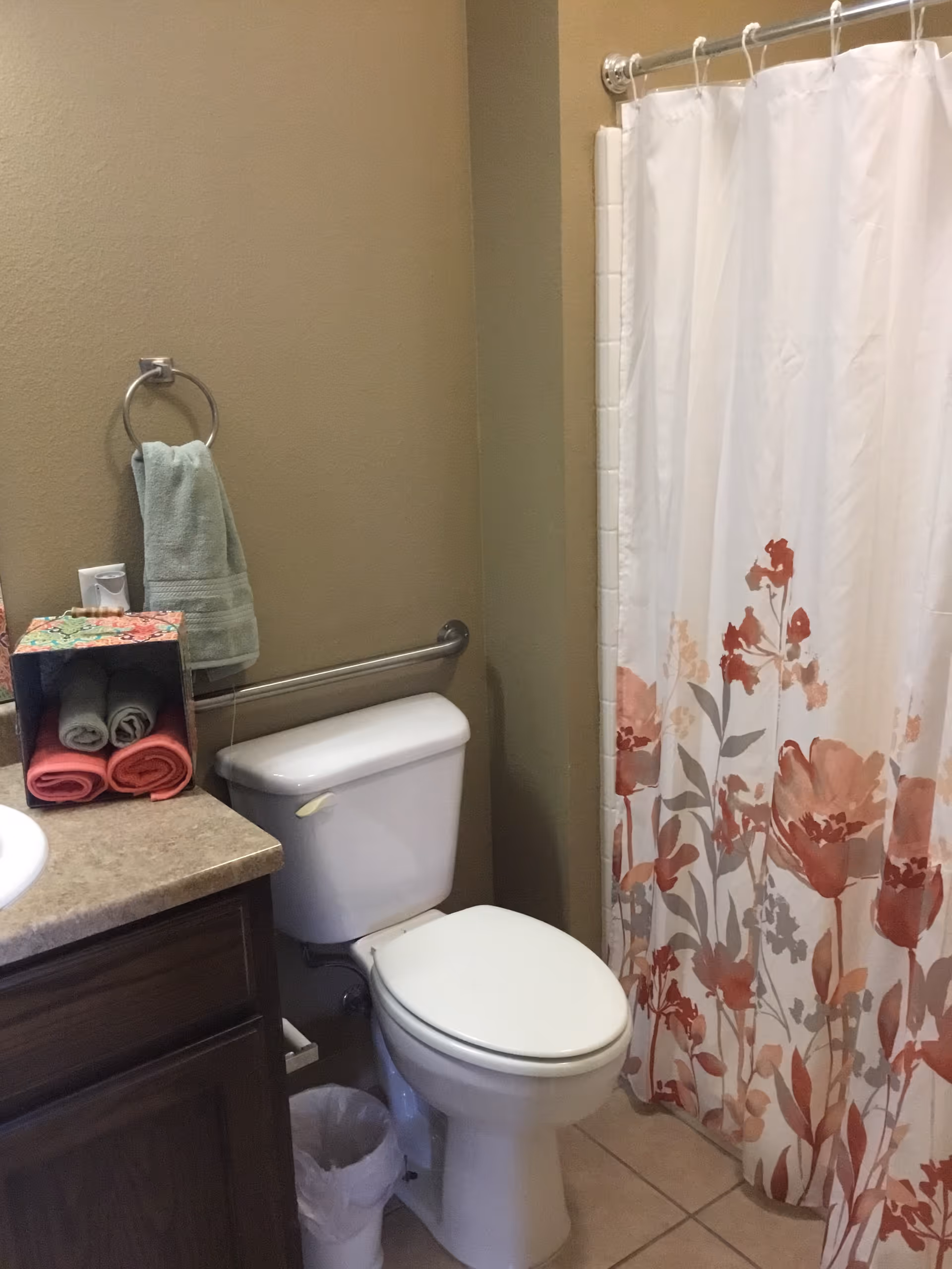 Bathroom with a toilet beside a sink countertop, a floral shower curtain, towel ring and folded towels.