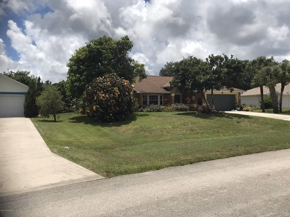 Single-story house with a green lawn, trees and a driveway under a cloudy sky.