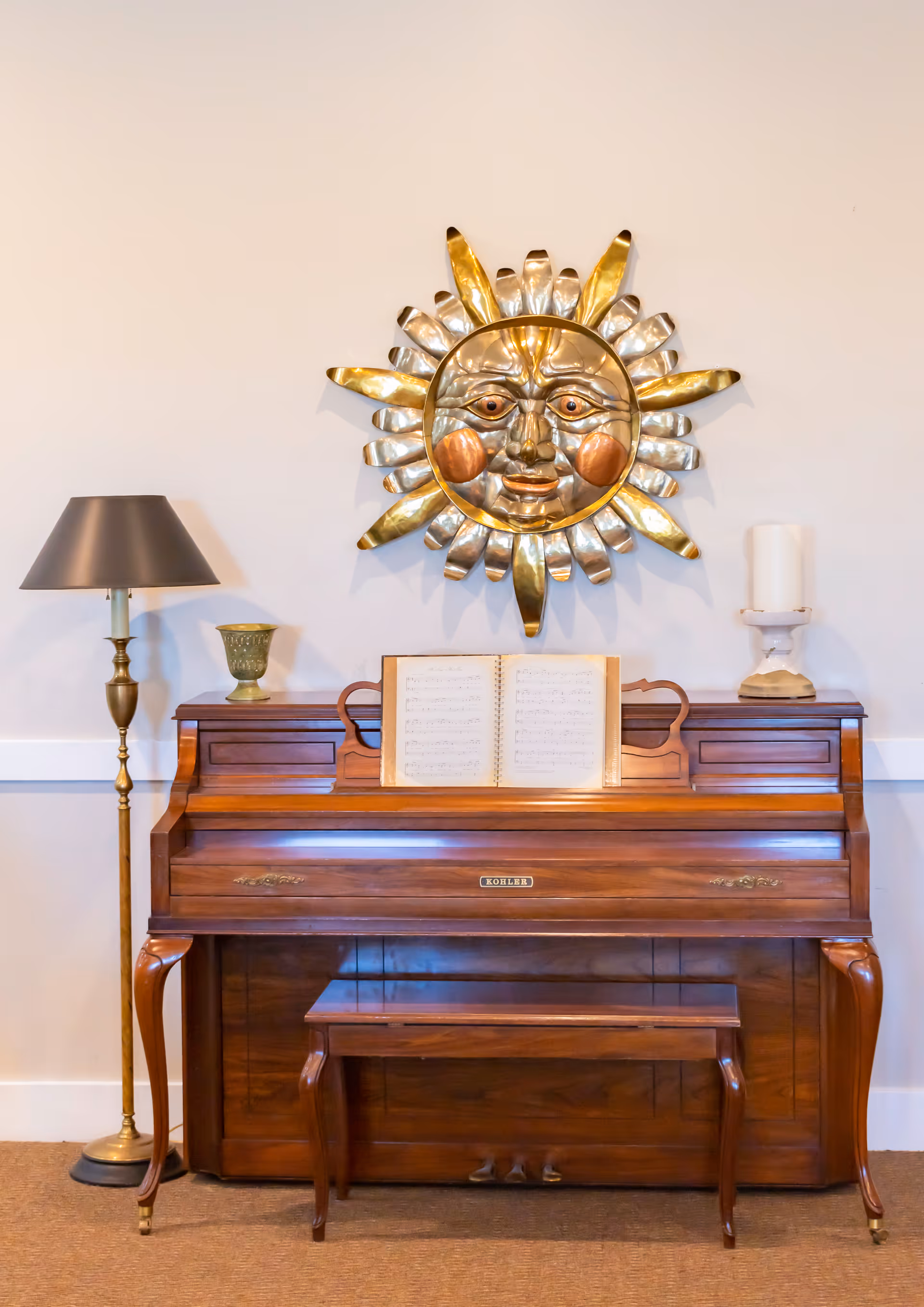 A wooden upright piano with a matching bench in front of a beige wall. On top of the piano is an open sheet music book, a small green decorative vase, and a white candle on a stand. Above the piano hangs a decorative metal sun with a face, featuring gold and silver tones. To the left of the piano is a tall brass floor lamp with a dark lampshade.