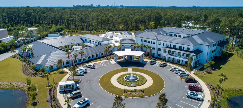 Aerial view of Watercrest Santa Rosa Beach senior living facility showing a large, modern building complex with multiple wings surrounding a circular driveway with a central fountain. Several cars are parked around the driveway, and the facility is surrounded by green lawns and trees.