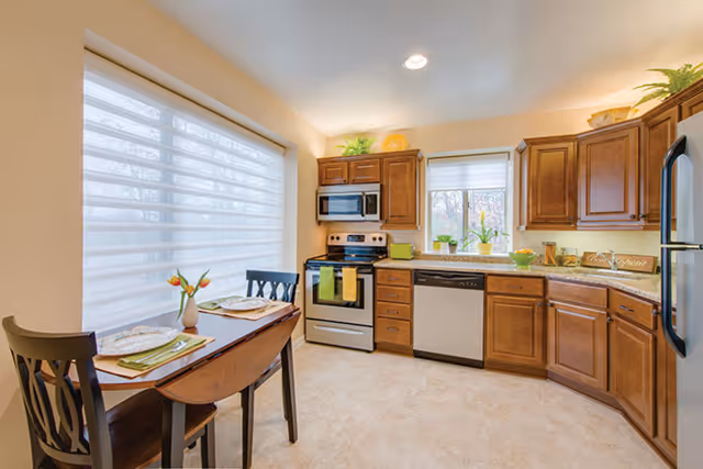 Well-lit kitchen with wooden cabinets, stainless steel appliances, a small dining table by a large window, and decorative plants.
