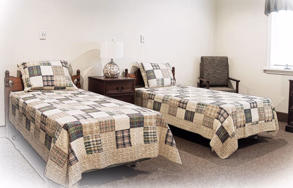 Two twin beds with patchwork quilts, a nightstand with a lamp, and a chair in a simple bedroom.