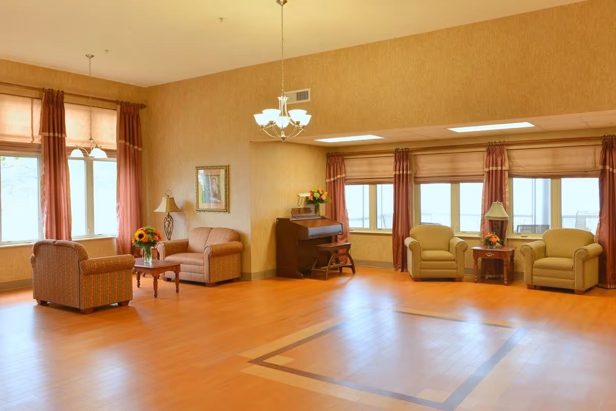 A spacious living room area with wooden flooring, beige walls, and large windows covered with brown curtains and blinds. The room features several upholstered armchairs and a loveseat arranged around small wooden tables with flower arrangements and lamps. A piano with a bench is positioned against one wall, and a chandelier hangs from the ceiling.